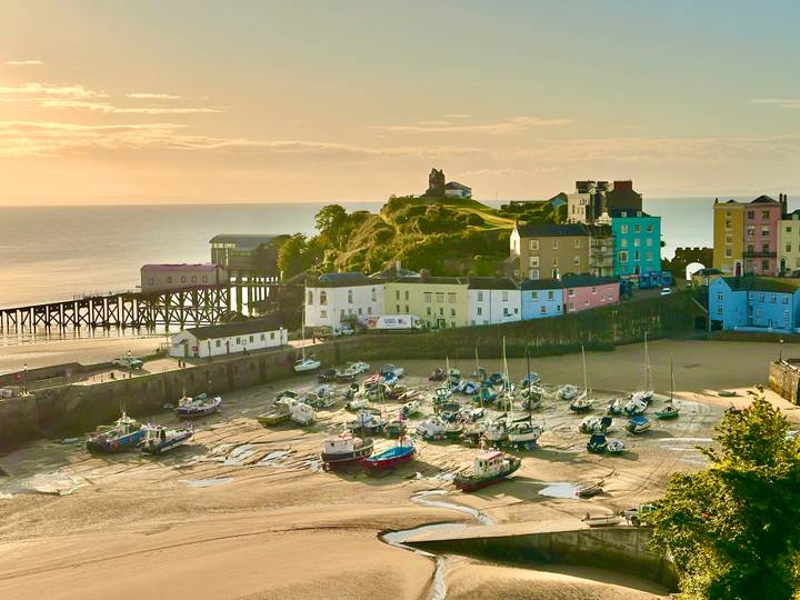 Golden evening light bathes Tenby harbour, colourful cliff-top houses and boats resting on tidal sand.