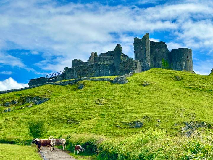 Ruined stone walls crown a grassy hill under a bright blue sky filled with wispy clouds.