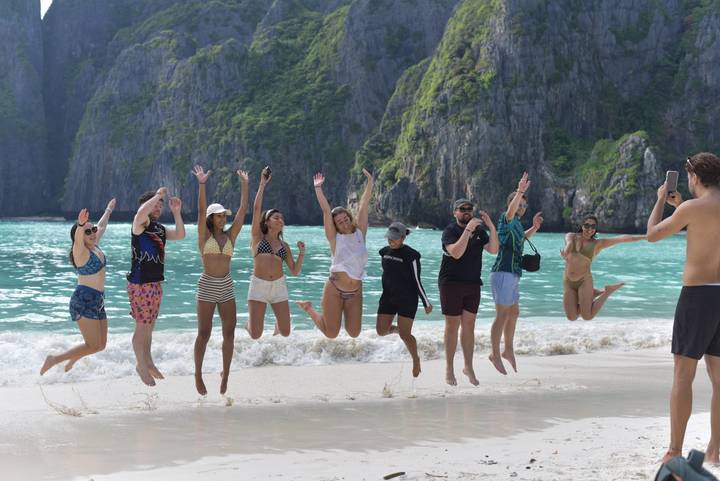 A lively group leaps simultaneously on a tropical beach backed by emerald cliffs and turquoise sea.
