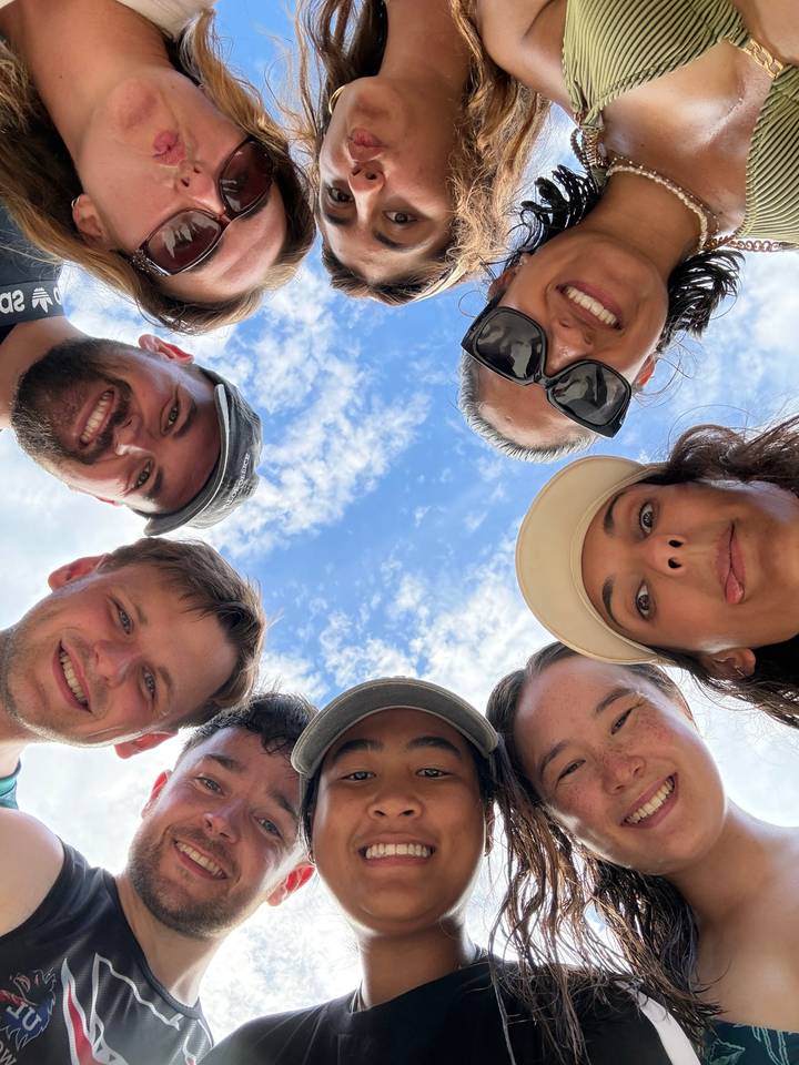 Friends huddle in a playful circle selfie, faces framed against a blue sky with scattered clouds.