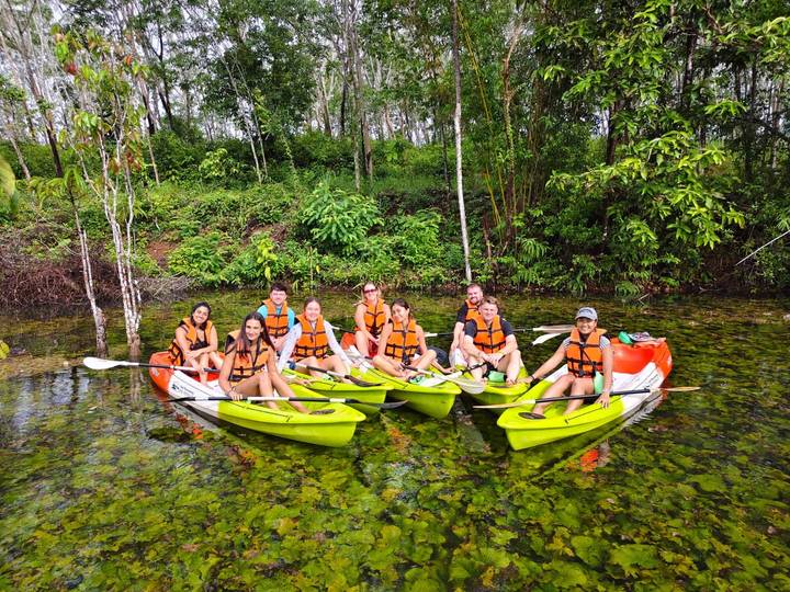A group in orange life vests sits in bright green kayaks on a calm jungle lagoon.