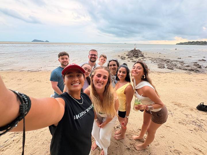 A cheerful group snaps a beach selfie with shallow tide flats and distant islands behind.
