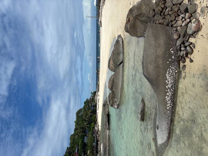 Clear shallows and smooth granite boulders line a quiet tropical beach under a partly cloudy sky.
