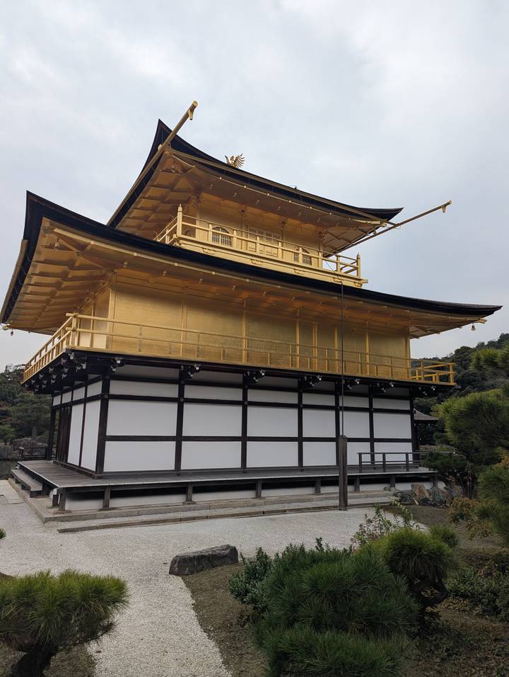 Side view of the gold-leaf-covered upper stories of Kinkaku-ji temple against a pale sky.