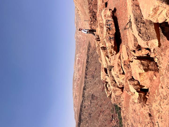 Lone traveler stands on red sandstone outcrop overlooking a vast arid plateau and blue sky.