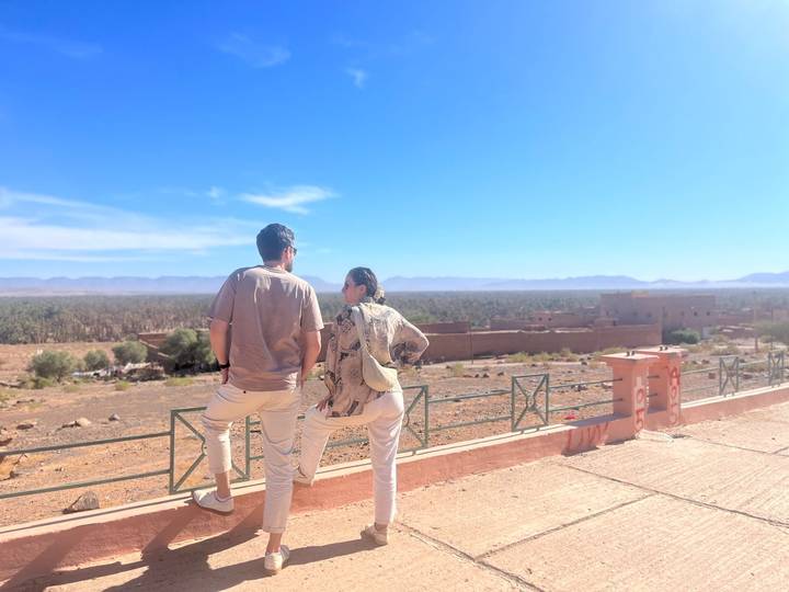 Couple leaning on a terrace rail gazing over a vast palm grove and desert plain.