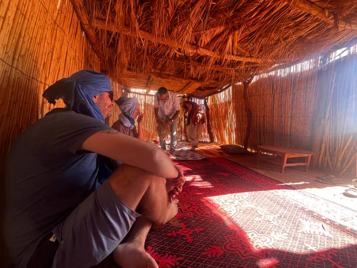 Travelers relax inside a thatched desert hut, sunlight filtering through woven walls.
