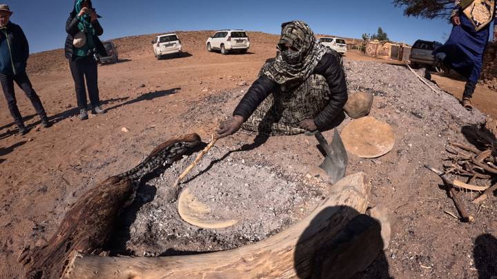 Guide in patterned scarf bakes flatbread in hot desert ashes as onlookers watch.