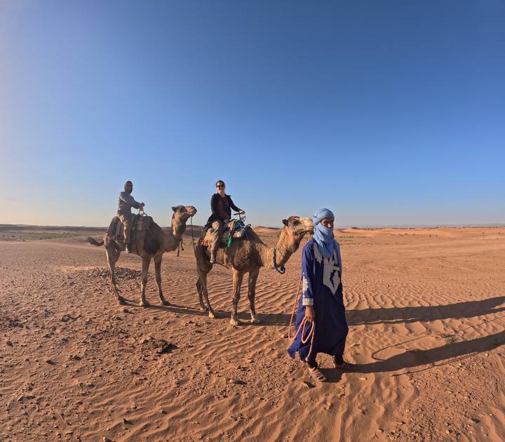 Camel caravan led by a blue-robed guide traverses rippled sand at golden hour.