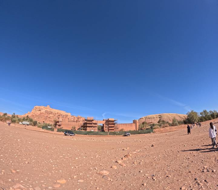 Panoramic shot of fortified clay buildings under scaffold near Ait Benhaddou with tourists scattered in foreground.