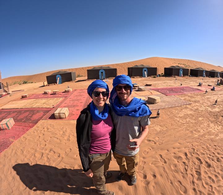 Smiling couple in blue turbans stand in front of luxury desert camp tents on soft sand.