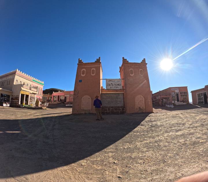 Traveler stands before twin adobe gate towers marking a desert town entrance under bright sun.