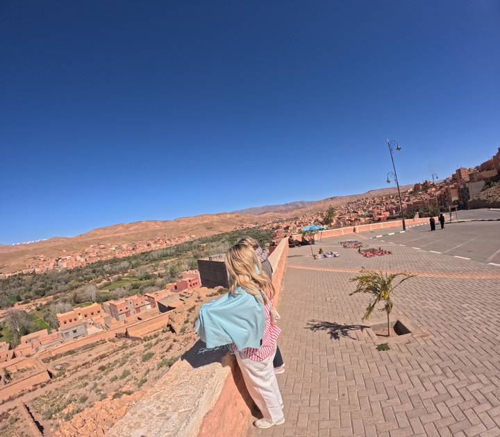 Woman with shawl looks out over terracotta town and arid hills from roadside terrace.