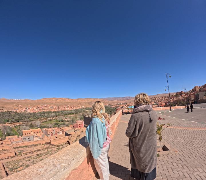 Two travelers admire a sprawling clay town and valley from a high viewpoint.
