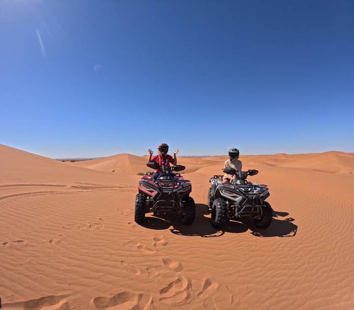 Two riders wearing helmets pose on quad bikes amid rolling orange sand dunes under a cloudless sky.