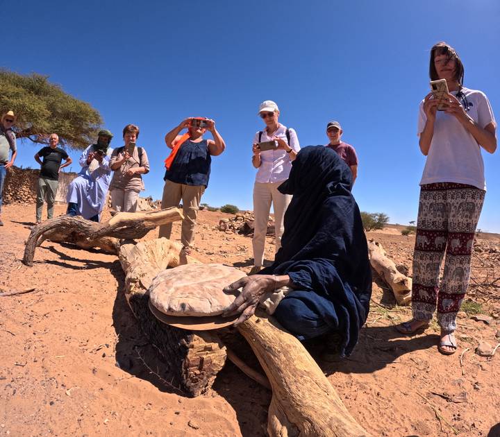 Travelers watch a veiled local woman bake flatbread over a stone in a sandy village clearing with a tree providing shade.