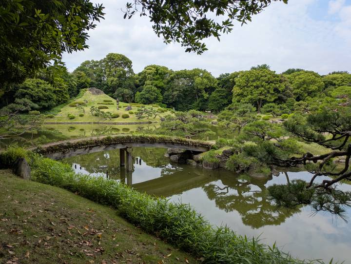 A curved wooden bridge crosses a reflective pond in a lush traditional Japanese garden.
