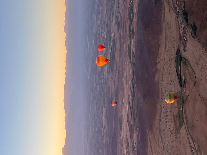 Multiple colorful hot-air balloons drift above a vast desert plain with distant mountain range at sunrise
