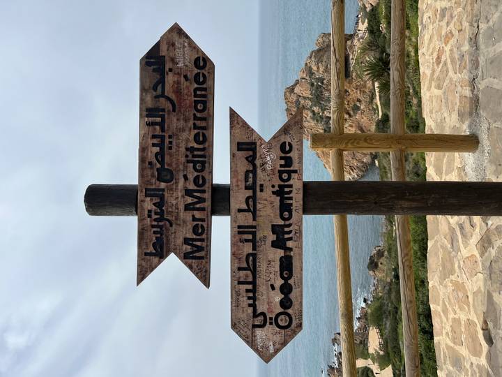 Bilingual wooden sign points toward the Mediterranean Sea and Atlantic Ocean with rocky coastline behind