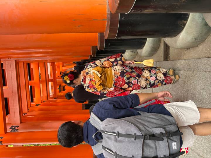 Visitors walk through a tunnel of vivid orange shrine gates; a woman in kimono leads the way.