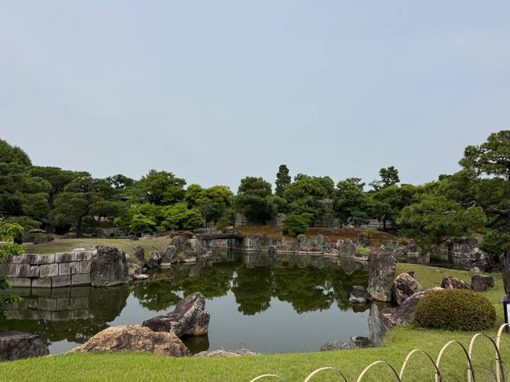 Serene Japanese garden pond reflecting scattered rocks and manicured greenery under a pale sky.