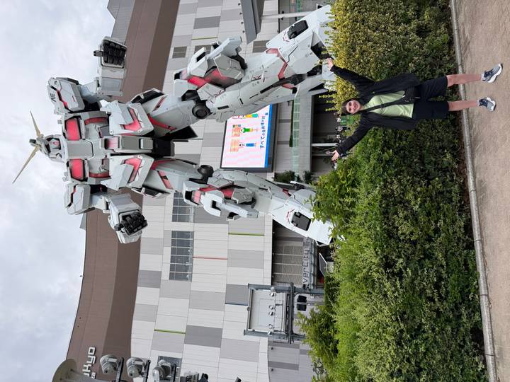 Traveller poses excitedly before a towering white Unicorn Gundam statue outside a mall.