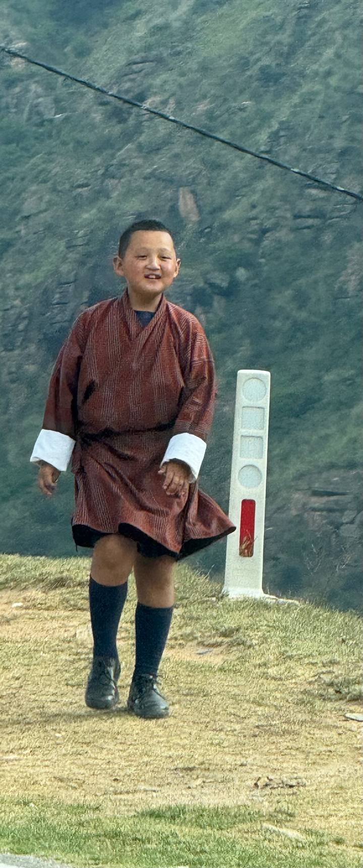 Bhutanese man in traditional gho walking beside a roadside marker against a misty mountain backdrop