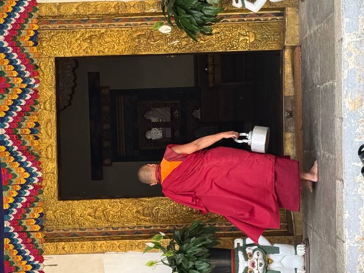 Buddhist monk in red robes carrying a kettle steps into a richly carved golden doorway