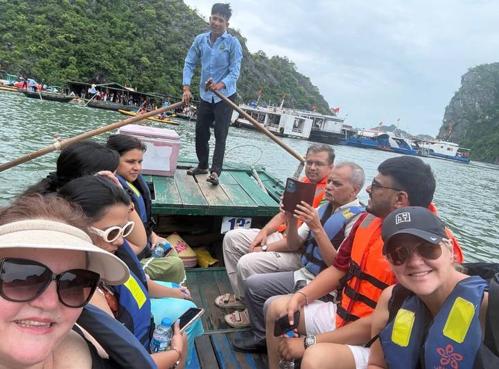 Tourists in life vests sit in a wooden boat rowed across emerald water with karst hills around.