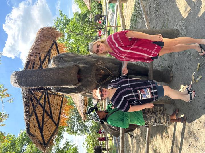Two women posing beside a large elephant reaching its trunk toward the camera under a thatched shelter.