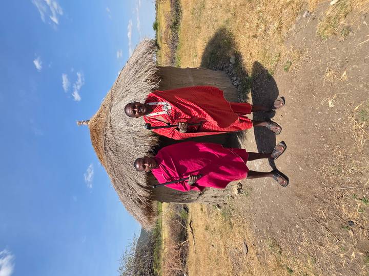 Two Maasai men in traditional red shukas stand outside a thatched hut.