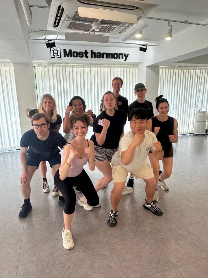 Energetic class of travelers pose in fighting stances inside a bright studio after a martial arts lesson.