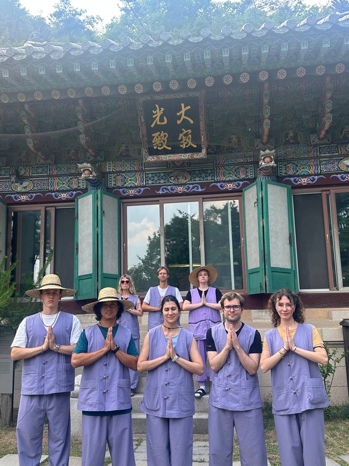 Group wearing traditional temple stay uniforms sit meditating in front of an ornately painted wooden building.