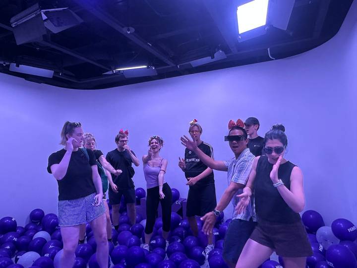 A group of young travelers dance and pose in a purple-lit room decorated with balloons and fun headbands.