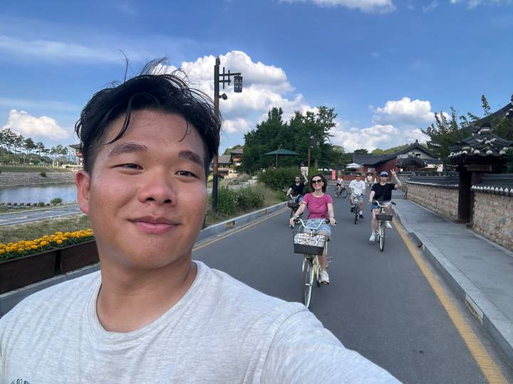 A smiling cyclist takes a selfie while friends ride bicycles down a traditional village street on a sunny day.