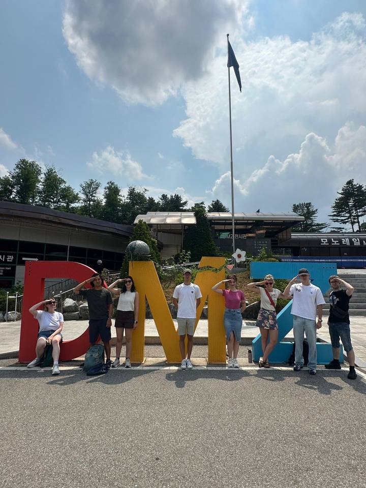 Travelers salute playfully in front of a colorful DMZ sign outside a visitor center on a bright day.