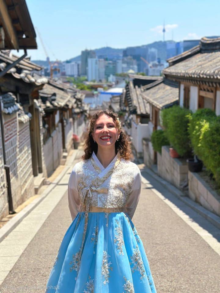 A woman wearing an elegant hanbok smiles in a sunlit historic alley lined with traditional wooden houses.