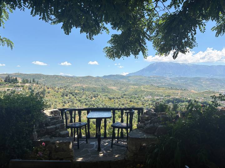 A shaded terrace with a table overlooks rolling valleys and distant mountains under a clear sky.