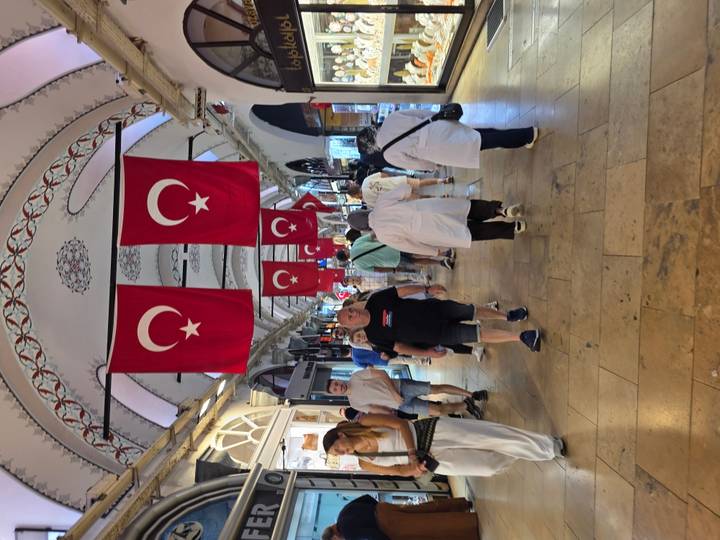 Crowd walking through Grand Bazaar corridor adorned with hanging Turkish flags.