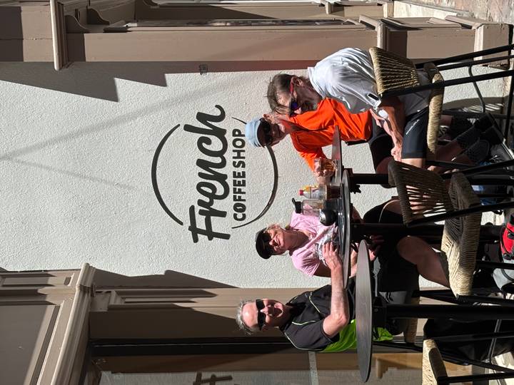 Cyclists relaxing with drinks at a sidewalk table outside a café that reads 'French Coffee Shop'.