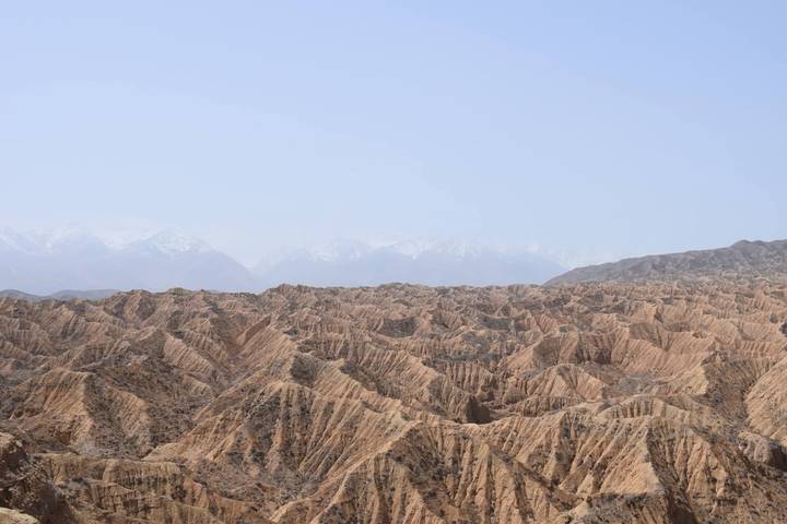 Vast badlands landscape with eroded ridges and distant snowy Tian Shan peaks under hazy sky