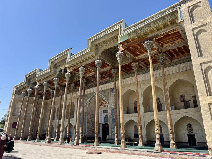 Wood-columned mosque façade with intricate ceiling carvings under clear sky