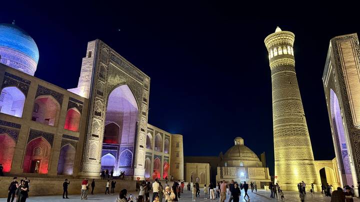 Night scene of Bukhara’s Po-i-Kalyan complex with the illuminated minaret and madrasah, visitors strolling below.