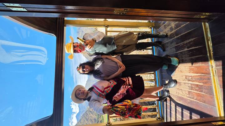 Three people, one in traditional costume, posing in the glass-roof observation car of an Andean train.