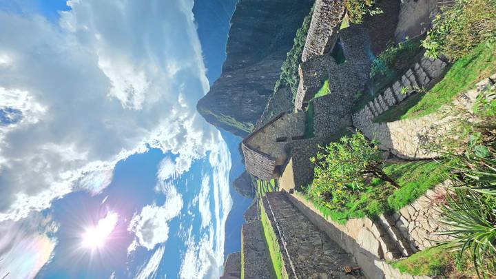 Sun-lit view of Machu Picchu terraces, stone houses and surrounding peaks beneath a dramatic sky.
