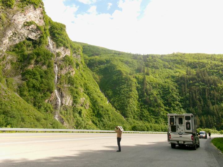 A lone traveller photographs a slender roadside waterfall cascading down lush green mountains beside a highway and camper van.