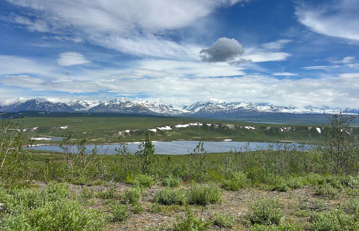 Wide Alaskan tundra with a small lake and endless snow-draped range beneath a dramatic cloud-streaked sky.