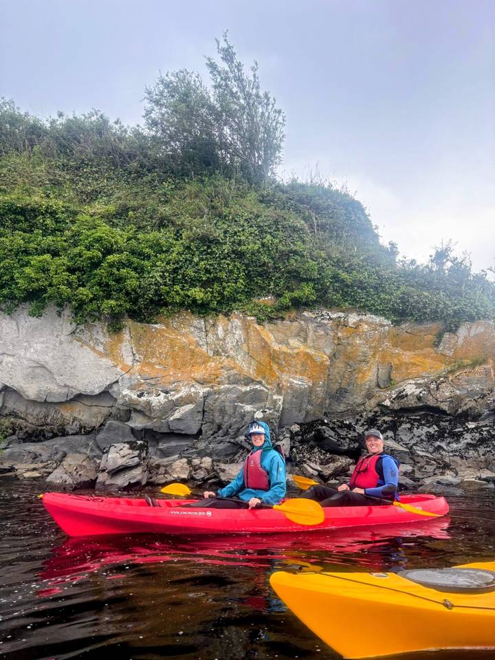 Two kayakers posing by a rocky, seaweed-covered shoreline beneath lush vegetation.