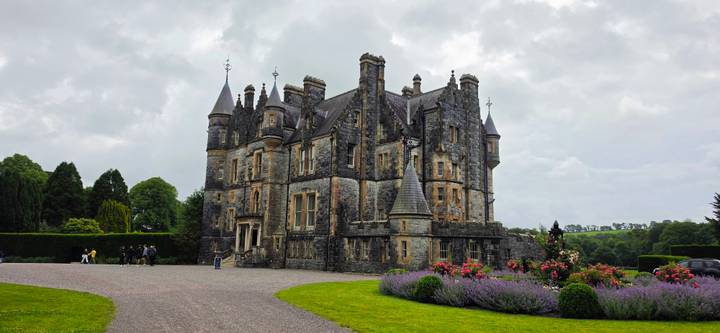 Impressive grey-stone neo-Gothic castle with turrets and gardens under a moody sky.