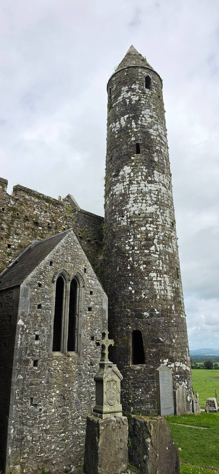 Ancient stone round tower and chapel ruins with weathered lichen under grey skies.